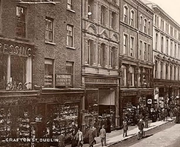 Black and white photograph of Grafton Street, Dublin, circa 1885, showing Victorian-era commercial buildings with ground-floor shops, pedestrians walking along the pavement, and a busy street scene