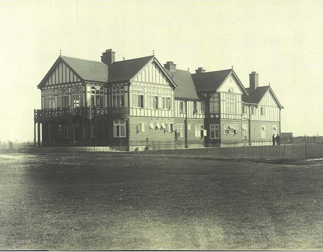 Early 20th-century black and white photograph of the original Golf Club of Dublin Tudor-style clubhouse at Dollymount, showing the timber-framed facade, first floor balcony, and iron perimeter fence viewed from the golf course
