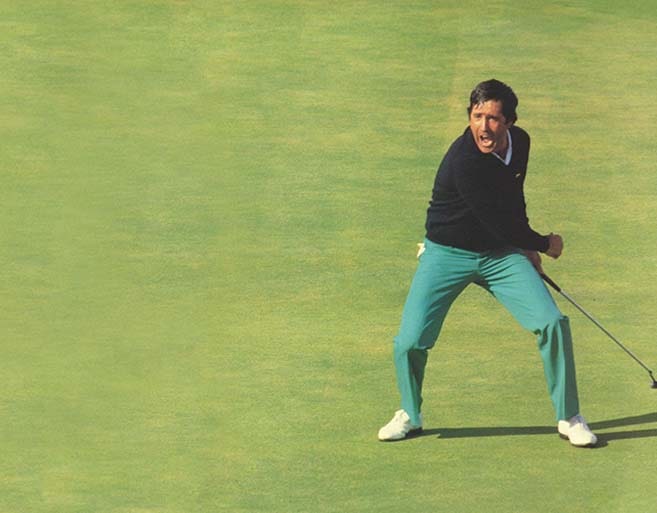 Colour photograph of Seve Ballesteros celebrating exuberantly on a golf green after holing a putt, wearing a navy sweater and teal trousers while holding his putter aloft