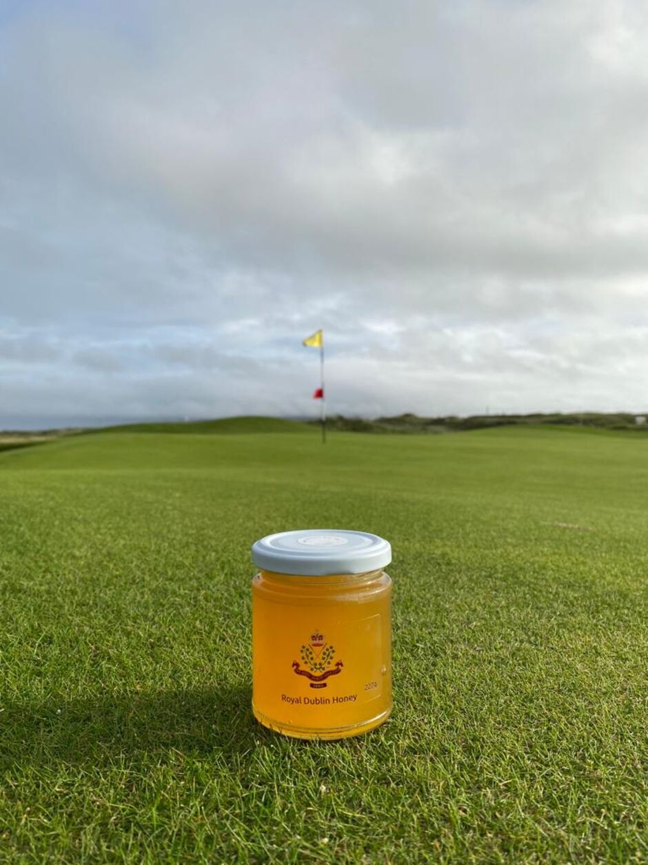 A small jar of Golf Club of Dublin Honey bearing the club crest, placed on the fairway grass at Golf Club of Dublin with a yellow and red pin flag on the green and a dramatic cloudy sky visible in the background