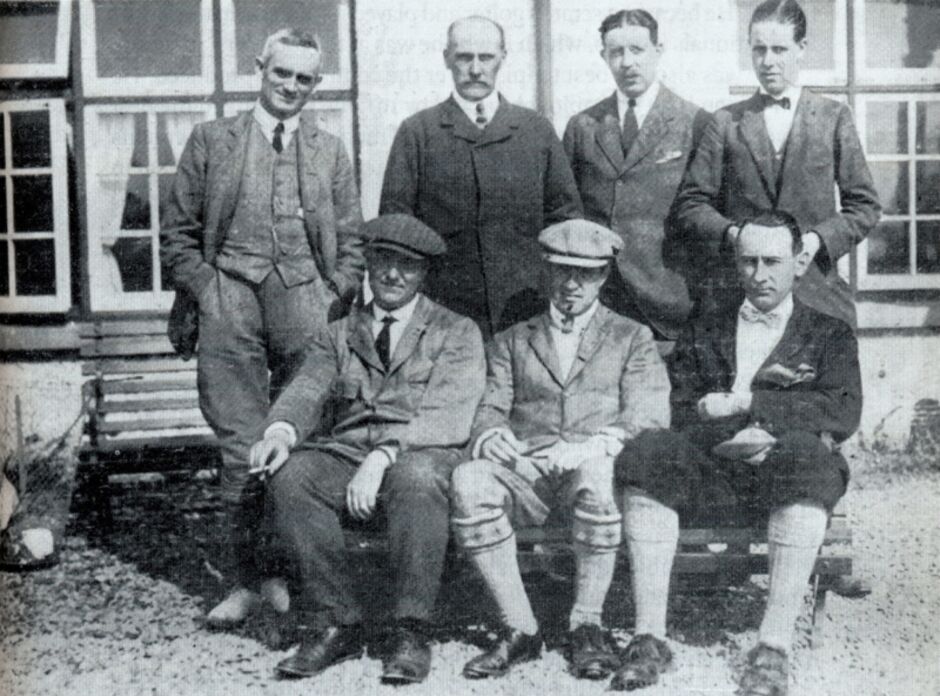 Black and white group photograph from 1914 of the Golf Club of Dublin Senior Cup team versus Portmarnock, showing seven men posed in two rows outside a clubhouse, three seated in front wearing plus-fours and flat caps, and four standing behind in suits