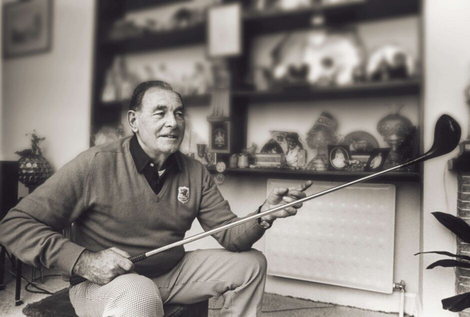 Black and white portrait photograph of Irish golfing legend Christy O'Connor Senior seated indoors holding a golf club, wearing a v-neck sweater with a club crest, with shelves of trophies and memorabilia visible in the background