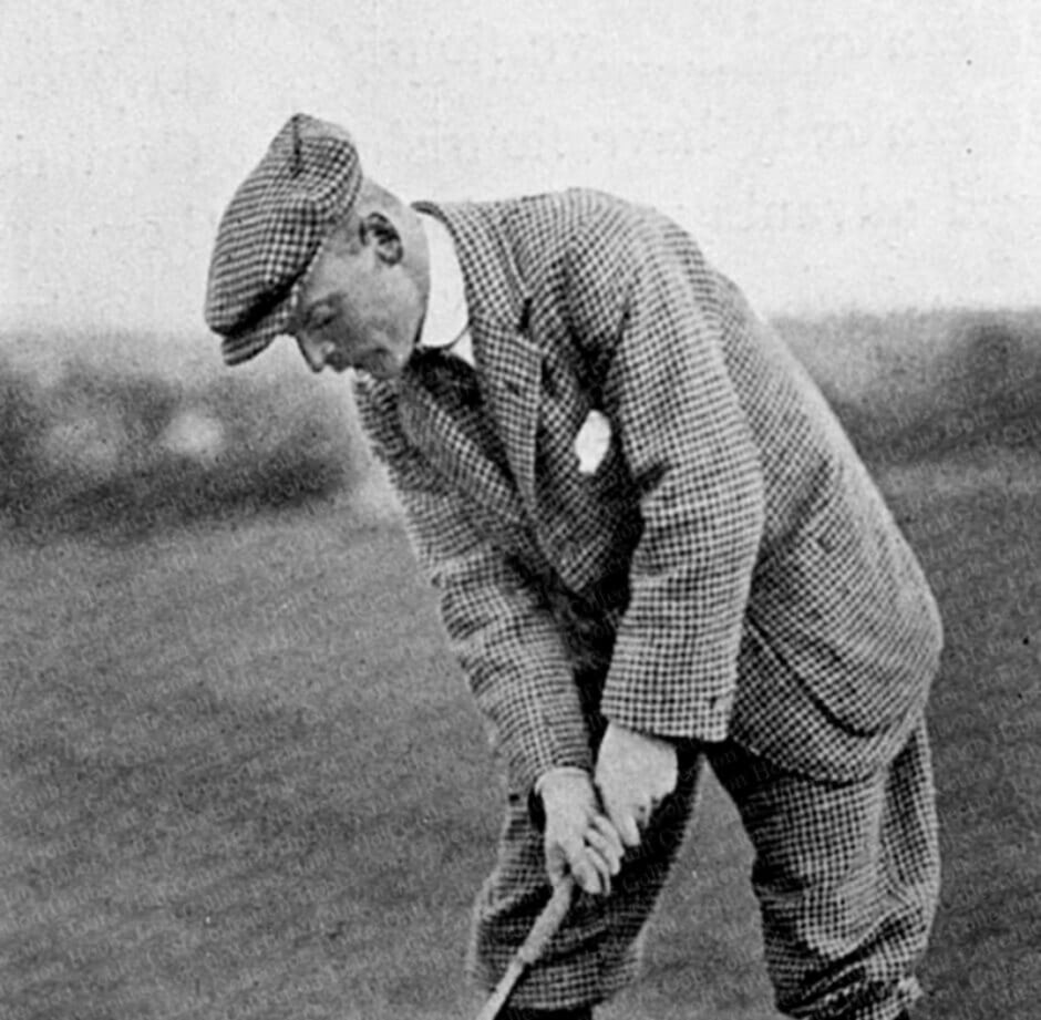 Black and white photograph circa 1910 of golf course designer Harry Shapland Colt addressing a golf ball, wearing a checked tweed suit and flat cap, shot on a links course