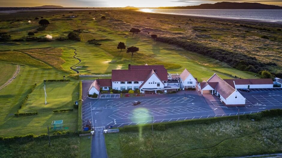 Aerial drone photograph of Golf Club of Dublin's white-rendered clubhouse with red roof at sunset, surrounded by the links golf course, car park, and Dublin Bay coastline with the Wicklow Mountains visible on the horizon